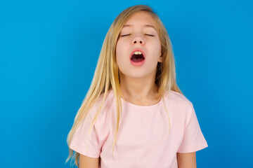 Caucasian kid girl wearing pink shirt against blue wall yawns with opened mouth stands. Daily...