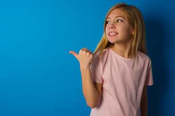 Caucasian kid girl wearing pink shirt against blue wall points away and gives advice demonstrates advertisement