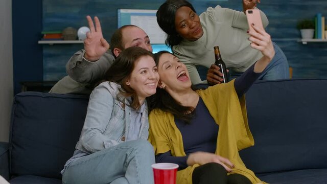 Group of mixed race people taking pictures with phone while sitting on couch in living room spending time together. Multiracial friends posting selfie on internet sharing with other person