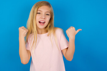 Fototapeta premium Hooray cool Caucasian kid girl wearing pink shirt against blue wall point back empty space hand fist