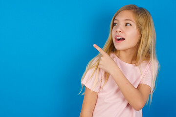 Caucasian kid girl wearing pink shirt against blue wall glad cheery demonstrating copy space look novelty