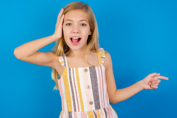 Surprised Caucasian kid girl wearing dress against blue wall pointing at empty space holding hand on head