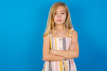 Confident Caucasian kid girl wearing dress against blue wall with arms crossed looking to the camera