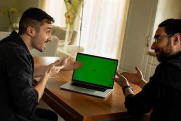 Zwei junge Männer, sehen ausländisch aus, sitzen im Wohnzimmer vom Laptop und haben eine Geschäftsidee. Einer zeigt mit der Hand auf dem PC, der andere zeigt begeistert auf green screen Bildschirm