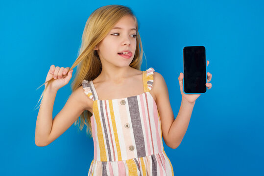 Photo Of Nice Pretty Caucasian Kid Girl Wearing Dress Against Blue Wall Demonstrate Phone Screen Hold Hair Tails