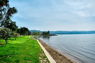 Fototapeta premium A shore and water's edge of Nicaea (iznik) lake during sunny day and with beach covered by green trees and grass with small pathway near it and small hill background and blue sky.