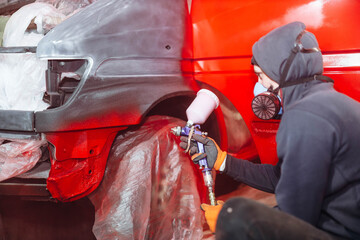 man in a respirator paints a car with a spray gun in red, repair a truck body.