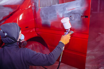 man in a respirator paints a car with a spray gun in red, repair a truck body.