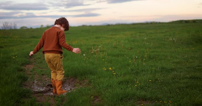 happy redhead boy, kid having fun on a spring meadow, jumping in muddy puddle