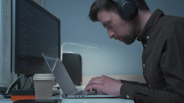 Handsome male software developer programming codes while working behind laptop with headphones from home. IT engineer in headphones working on computer in office. Freelancer works, social distancing