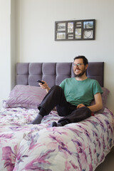 Happy man zapping and watching tv relaxing in the bed smiling. He is wearing green t-shirt and black pants.	
