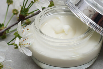 Beauty cream in a glass jar on a light gray background. Decorated with white spring flowers. Unbranded skincare product. Cosmetic cream. Close up, selective focus. - 431817726
