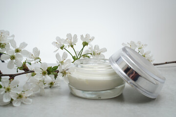 .Beauty cream in a glass jar on a light gray background. Decorated with white spring flowers. Unbranded skincare product. Cosmetic cream. Close up, selective focus, side view. - 431817744