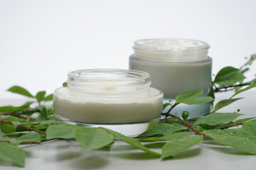 Glass jars with beauty cream on a light gray background. Decorated with green leaves. Unbranded skincare product. Cosmetic cream. Close up, selective focus, side view. - 431817751