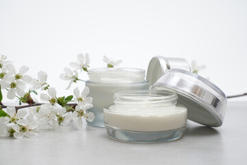 Beauty cream in a glass jars on a light gray background. Decorated with white spring flowers. Unbranded skincare product. Cosmetic cream. Close up, selective focus, side view. - 431817752
