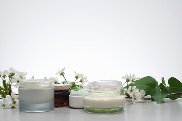 Beauty cream in a glass jars on a light gray background. Decorated with white spring flowers. Unbranded skincare product. Cosmetic cream. Close up, selective focus, side view. - 431817757