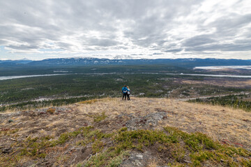 Two people, couple hikers standing on a panoramic view above Yukon River outside of Whitehorse in north of Canada. Boreal forest, snow capped mountains on a cloudy afternoon in distance. 