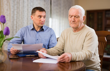 Troubled senior man discussing documents with his adult son while sitting at home table