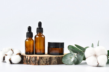 A brown glass bottle on a wooden podium and eucalyptus with cotton on a light blue background. Eco-friendly organic natural cosmetics for skin and body care. Copy space