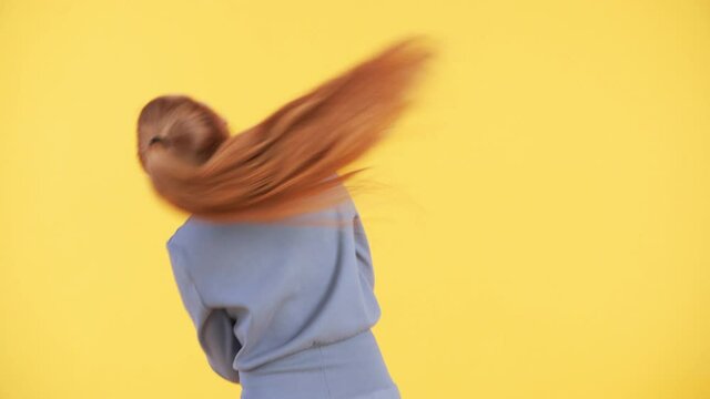 Back of a woman with long ginger ponytail dancing and moving her hair. Yellow background. Woman is dancing enjoying herself
