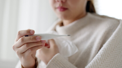 Naklejka premium Sick young asian woman sit under blanket on sofa and sneeze with tissue paper at home. Female blow nose, coughing, sneezing, check temp in tissue at home, suffering from flu. Cold and fever concept