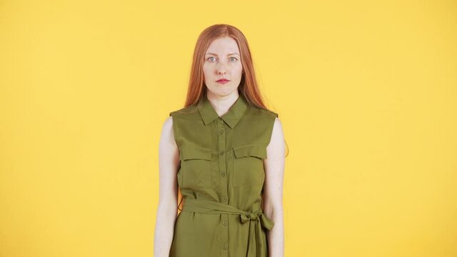 Young woman with freckles and long ginger hair is looking into the camera, "zipping" her mouth and walking away from the frame. Yellow background