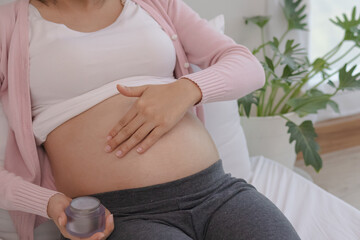Asain young pregnant woman applying pregnancy lotion to prevent stretch marks on belly by rubbing a cream with a moisturizer for abdomen beauty.