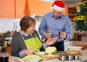 Senior woman cutting cabbage for salad in kitchen, man with glass of champagne