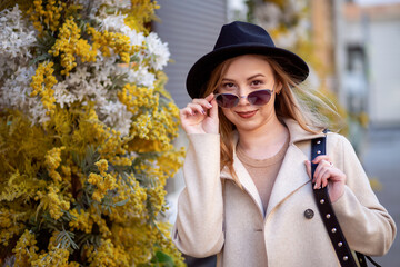 A young girl in sunglasses and a hat stands next to flowers - obrazy, fototapety, plakaty