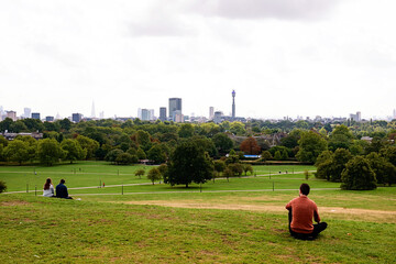 Primrose Hill in London with people sitting on grass. City skyline in misty day.