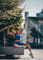 Slim athletic woman in sportswear, runner sits on bench form in city downtown, Dubai after workout exercises, looks aside, fixing hairstyle. Full-growth. Copy space
