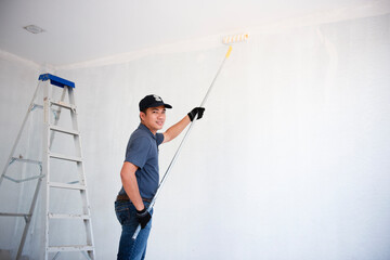 The back view of an Asian painter paints the walls with a paint roller and a separate bucket on a large empty space with a wooden staircase Young worker painting wall in room