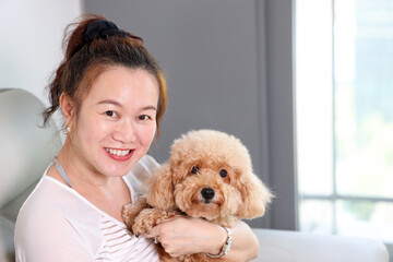 Beautiful attractive middle age southeast Asian woman sitting on sofa posing happy with small pet brown dog