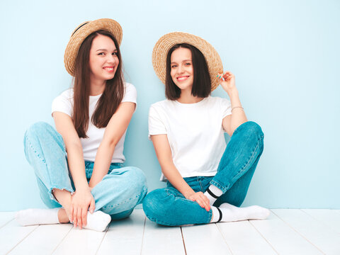 Two young beautiful smiling hipster female in trendy summer white t-shirt and jeans clothes.Sexy carefree women posing near light blue wall in studio.Positive models in hats