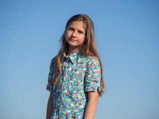 Fototapeta premium A teenage girl on a blue background. A happy, young, beautiful blonde looks at the camera in a summer dress with flowers on a blue sky background.