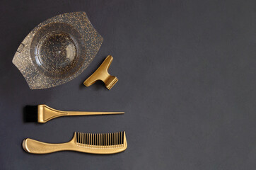 Golden hairdressing tools for hair coloring on a dark gray background with copy space. On the table are the accessories of the hair salon comb, brush, bowl and clips, top view.