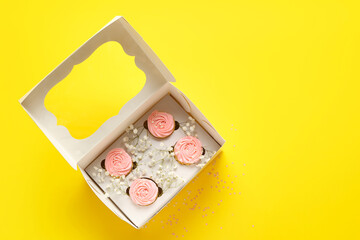 Box with tasty cupcakes and flowers on color background