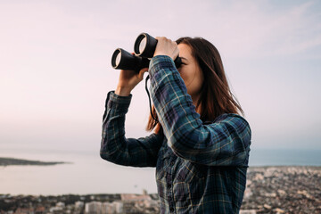 Portrait of a young female tourist looking through binoculars at sunset.