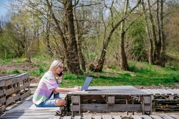 Fototapeta premium Blonde woman works outdoors using laptop and cell phone in beautiful sunny weather.