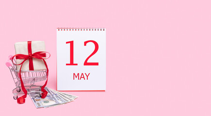 A gift box in a shopping trolley, dollars and a calendar with the date of 12 may on a pink background.