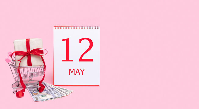 A gift box in a shopping trolley, dollars and a calendar with the date of 12 may on a pink background.