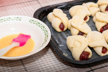 homemade cakes with jam on a baking sheet prepared for baking next to a plate of egg yolk for greasing