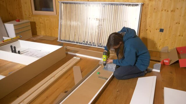 CLOSE UP: Caucasian female is screwing together metal bits and plywood boards while furnishing the bedroom of her brand new home. Young woman in blue hoodie assembles furniture with a power drill.