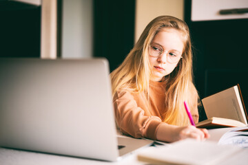 Beautiful 10 years old blond girl in glasses sitting at the desk doing her homework using a laptop.