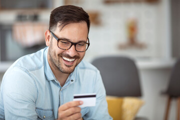Smiling relaxed man sitting on sofa shopping online using smartphone and credit card at home. Online payment. Modern businessman using laptop and credit card