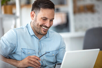 Young man working at home in the evening. Young businessman working at home using lap top. Young attractive smiling guy is browsing at his laptop, sitting at home