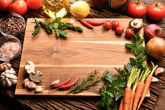 Empty wooden cutting board with copy space surrounded by peppers, herbs, tomatoes, mushrooms and other delicious flavored vegetables on table