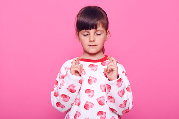 Charming concentrated female kid wearing casual jumper with hearts keeping eyes closed and fingers crossed, hopes for better, wish dream comes true, isolated over pink background.