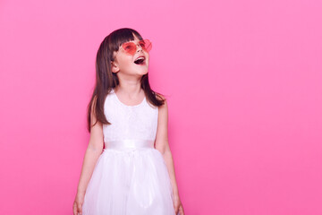 Cute little girl wearing fashionable glasses in heart shape and white dress, having excited facial expression, looking up with opened mouth and surprised look, sees something amazing.