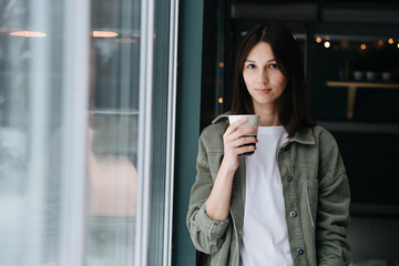 Attractive beautiful middle aged woman standing in a bedroom next to a window with mug in a hand. She's wearing casual clothes.